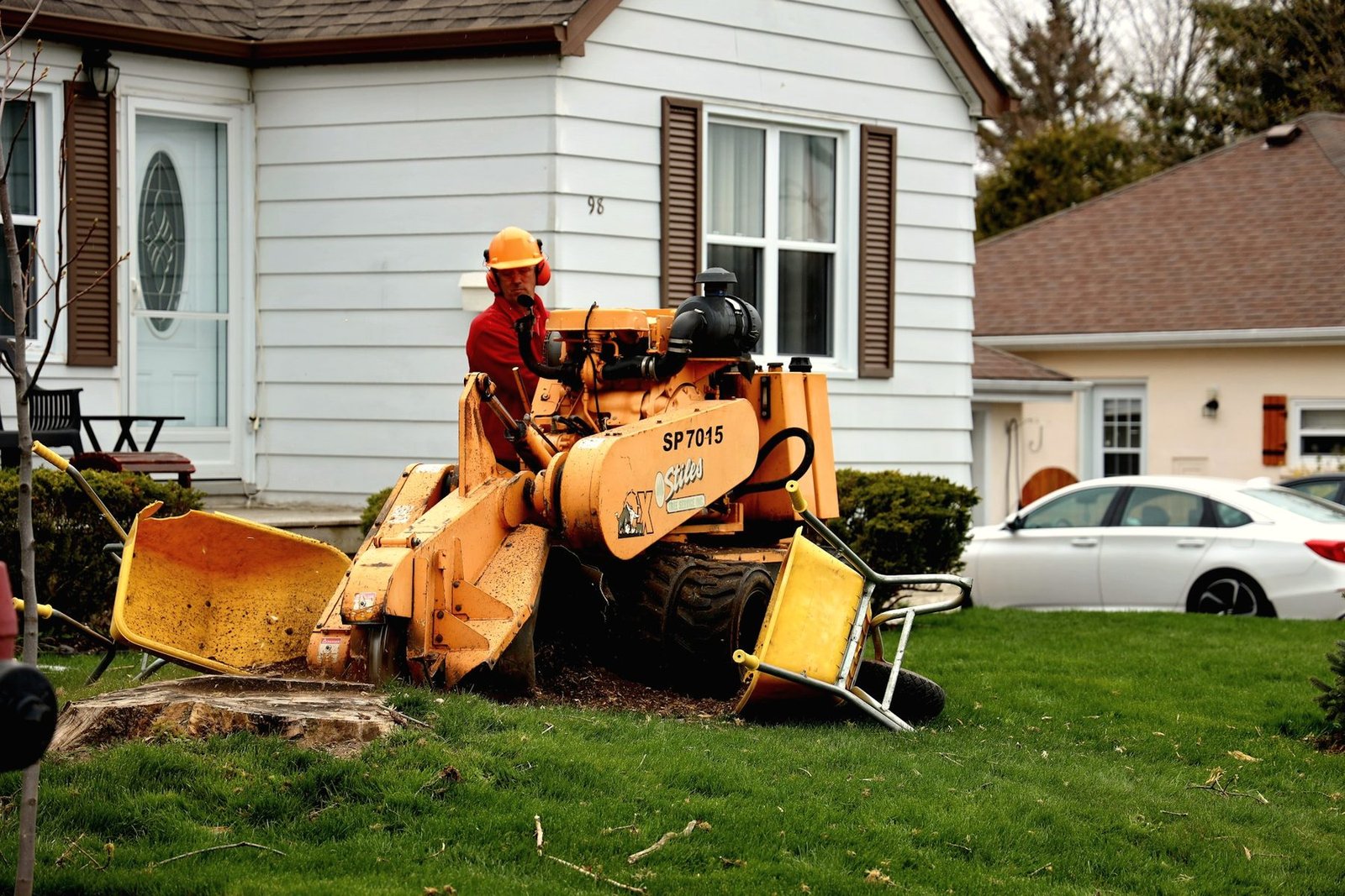 A worker in ear defenders running a stump grinder on a front lawn beside a white house